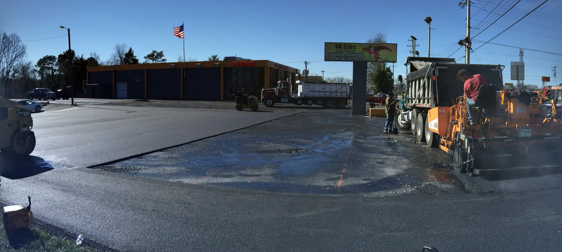 Paver and rollers working a dealership lot, business open behind the cones